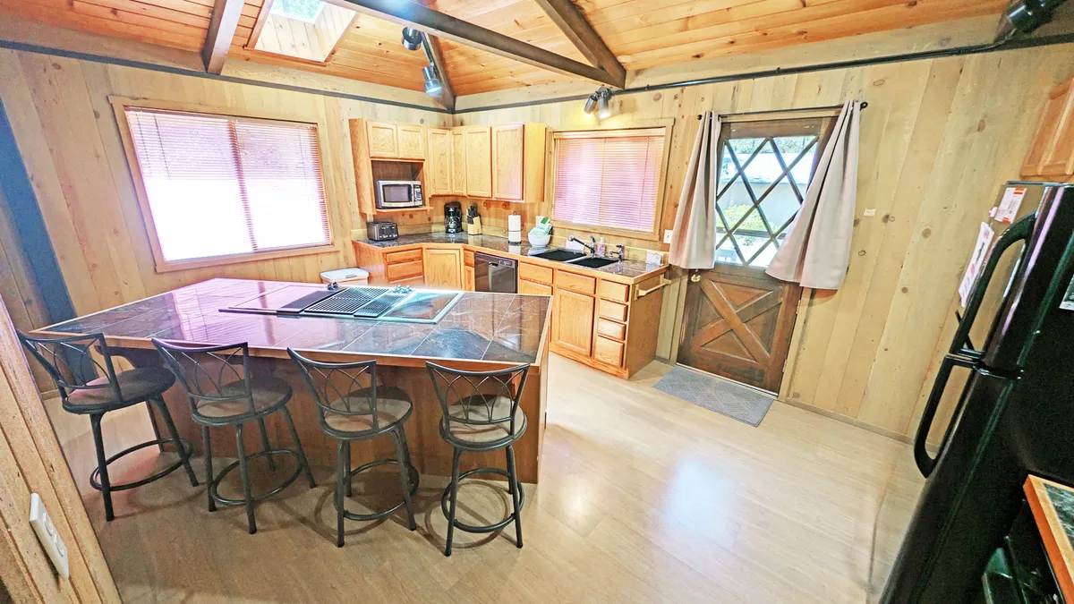 This image captures a warm kitchen and dining area with a large window and a door leading directly outside. The kitchen island and wooden cabinetry create a welcoming atmosphere, while the visible door-to-outside hints at easy access to outdoor adventures—perfect for coming and going with pets after a walk or bike ride.