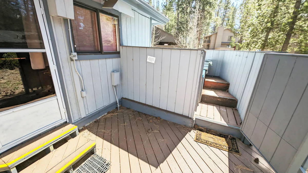 A wooden deck with steps leading down into a lush grove of tall trees. The deck features a welcome mat at the entrance, providing an inviting outdoor space for gathering, relaxing, or enjoying meals amidst the natural surroundings. The scene is framed by forest greenery, suggesting easy access to outdoor adventures and a strong connection to nature.