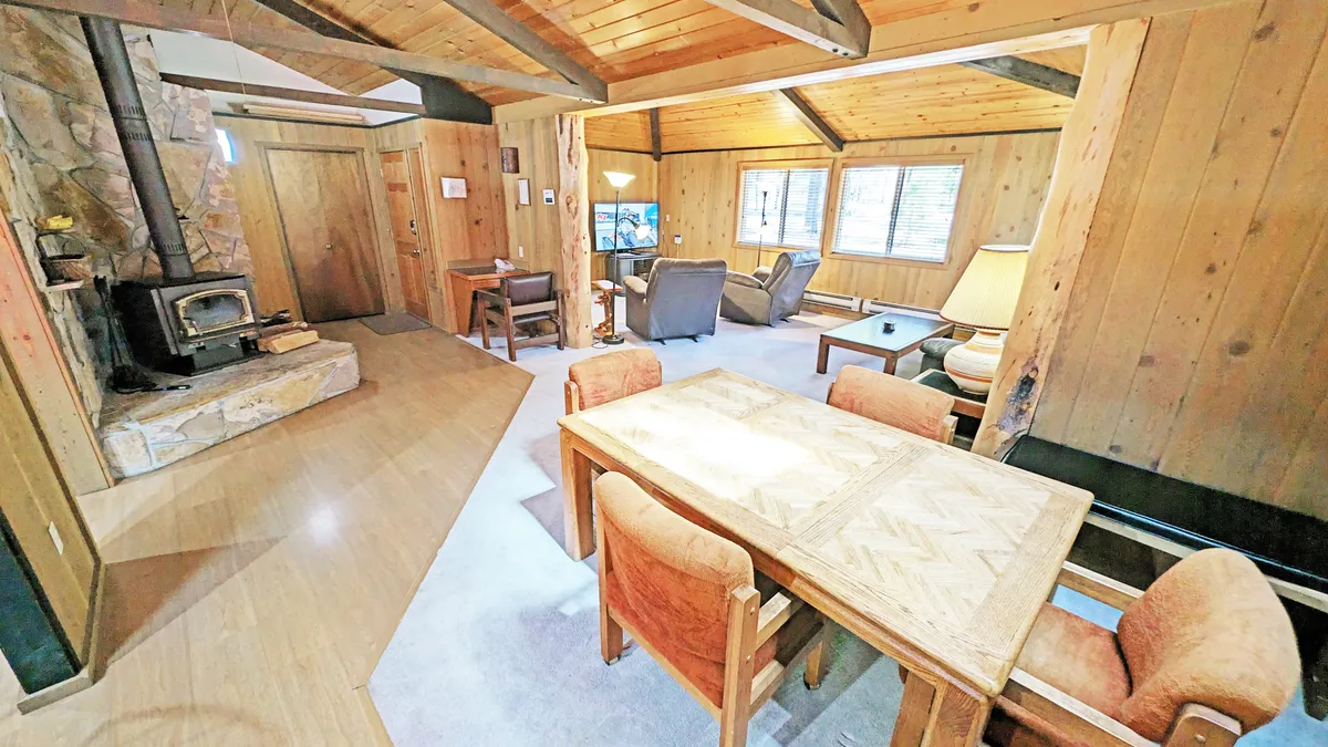 This image captures a warmly lit living area featuring a wood-burning stove, a comfortable seating area with plush sofas and chairs, a dining table, and expansive windows letting in natural light. The wood-paneled walls and exposed beams create a classic lodge atmosphere, and the arrangement encourages relaxation and gathering.