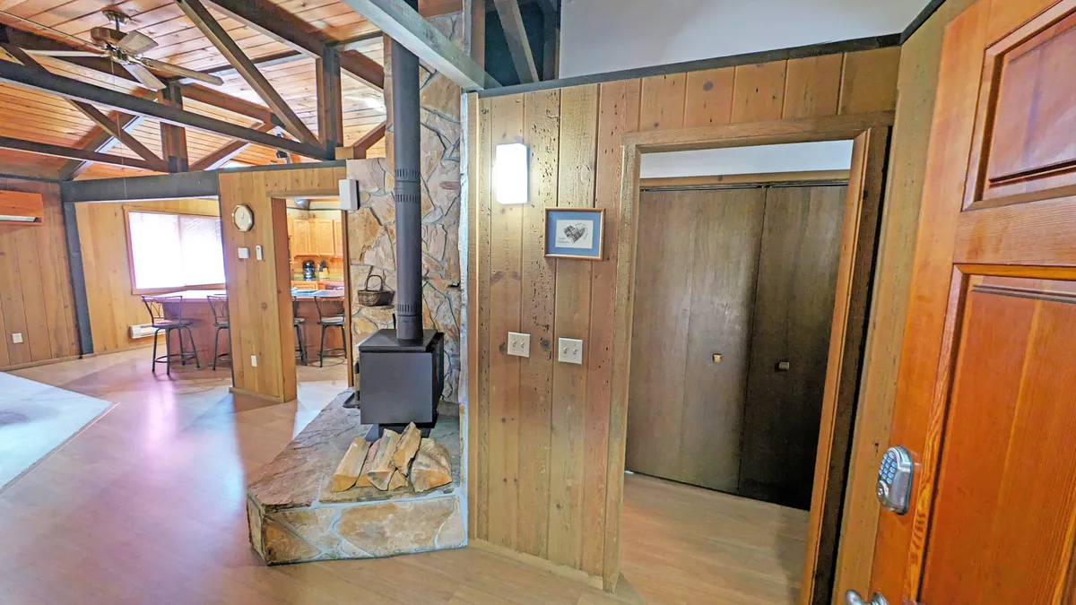 A wide-angle view of the open-concept living room, showcasing impressive wood-beamed ceilings, a stone fireplace (wood-burning stove style), and a welcoming seating area. The doorway to the kitchen hints at the flowing, communal layout. Large windows offer glimpses of the surrounding forest, bringing the outdoors in.