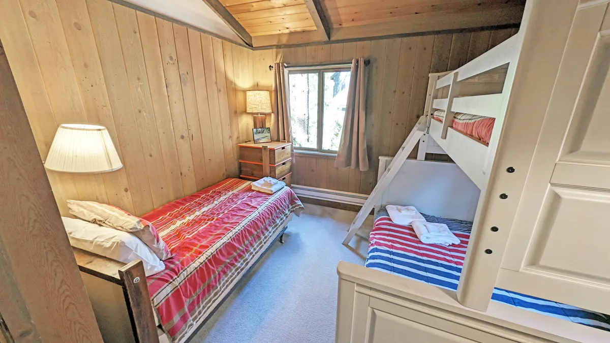 A cheerful guest bedroom with a pyramid-style bunk bed and a single twin bed. The room is bathed in natural light from a large window, and a lamp and nightstand add practical touches for guests, making it inviting for children or friends.