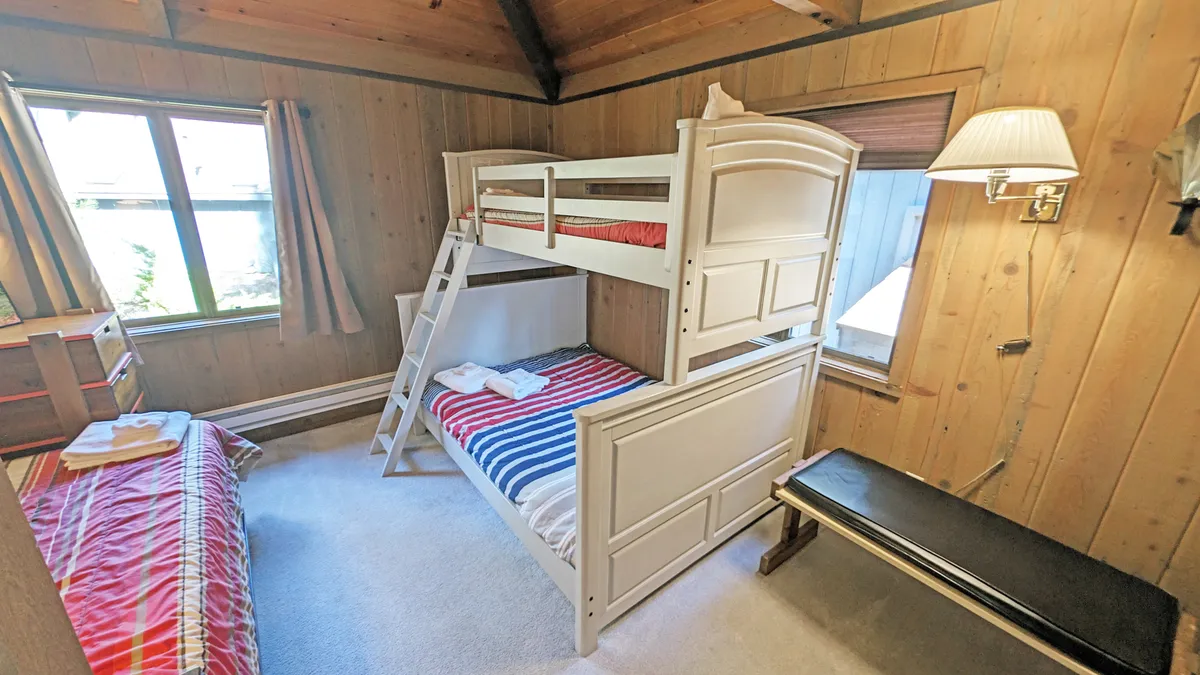 A close-up of a charming bunk bed with striped bedding against wooden walls, with a window letting in morning light and curtains adding a homey touch. The space feels inviting and playful—ideal for late-night chats or morning games.