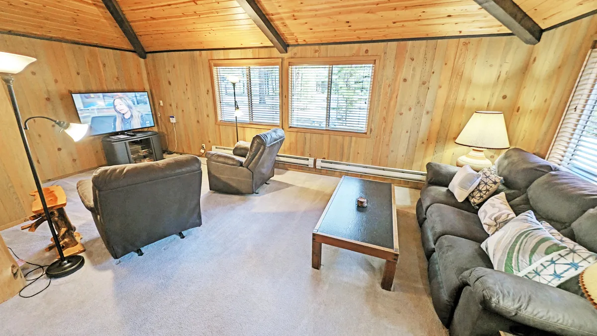 The image showcases a welcoming living room with plush sofas and recliners arranged around a coffee table and a television. The space features rich wood paneling, vaulted ceilings with large wood beams, and abundant natural light, creating a warm, cozy cabin atmosphere ideal for relaxing or gathering with loved ones.
