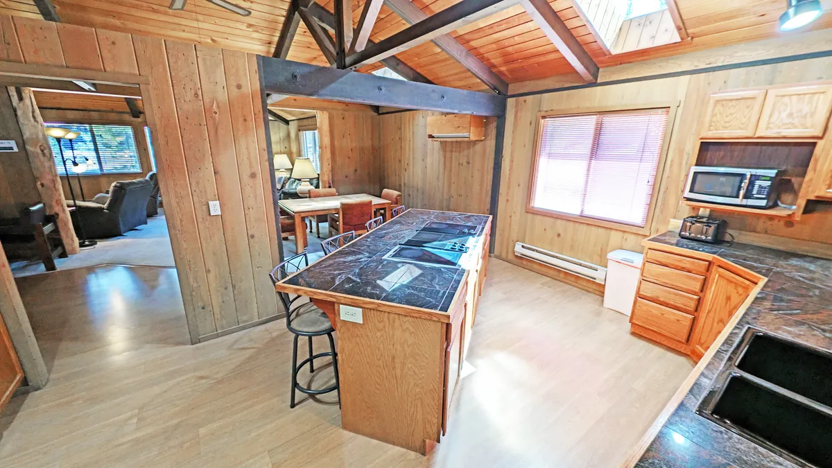 A sunlit kitchen scene with a kitchen island, exposed wooden beams, and a visible dining area alongside a large window. The overall feel is bright and airy, with the space inviting for family breakfasts, casual coffee breaks, or relaxed evening gatherings. The island acts as the heart of the kitchen, with bar stools suggesting opportunities for casual dining or conversation.