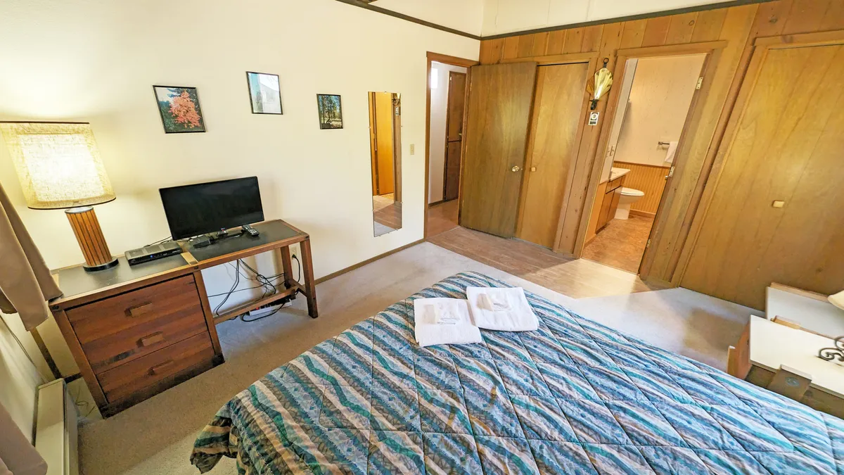 A cozy bedroom featuring a bed with a blue patterned quilt, a nightstand with a lamp, and a television on the desk. The room feels peaceful and private, with soft lighting that invites relaxation.