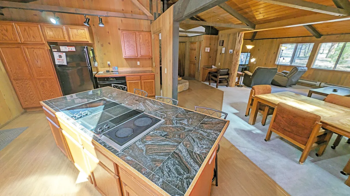 This image presents a modern, cozy kitchen with a prominent kitchen island, stainless steel appliances, warm wooden cabinetry, and a peek into the connected living area. There are bar stools at the kitchen island, emphasizing a communal space great for meal prep, snacks, or socializing. The overall mood is communal and inviting, perfect for group gatherings or family cooking sessions.